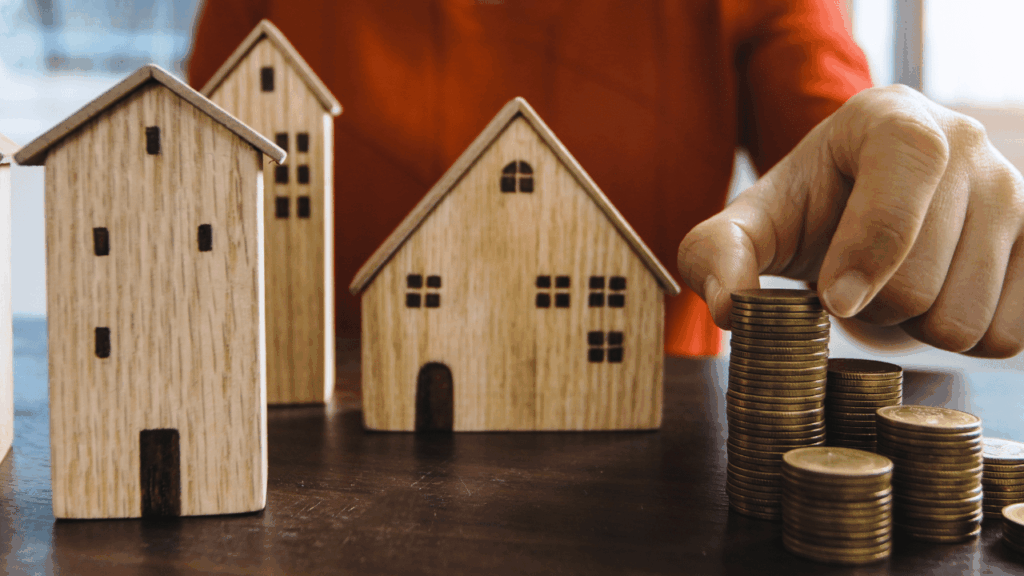 main plaçant une pièce sur une pile de pièces à côté de maquettes de maisons en bois, investissement immobilier, épargne pour achat maison.